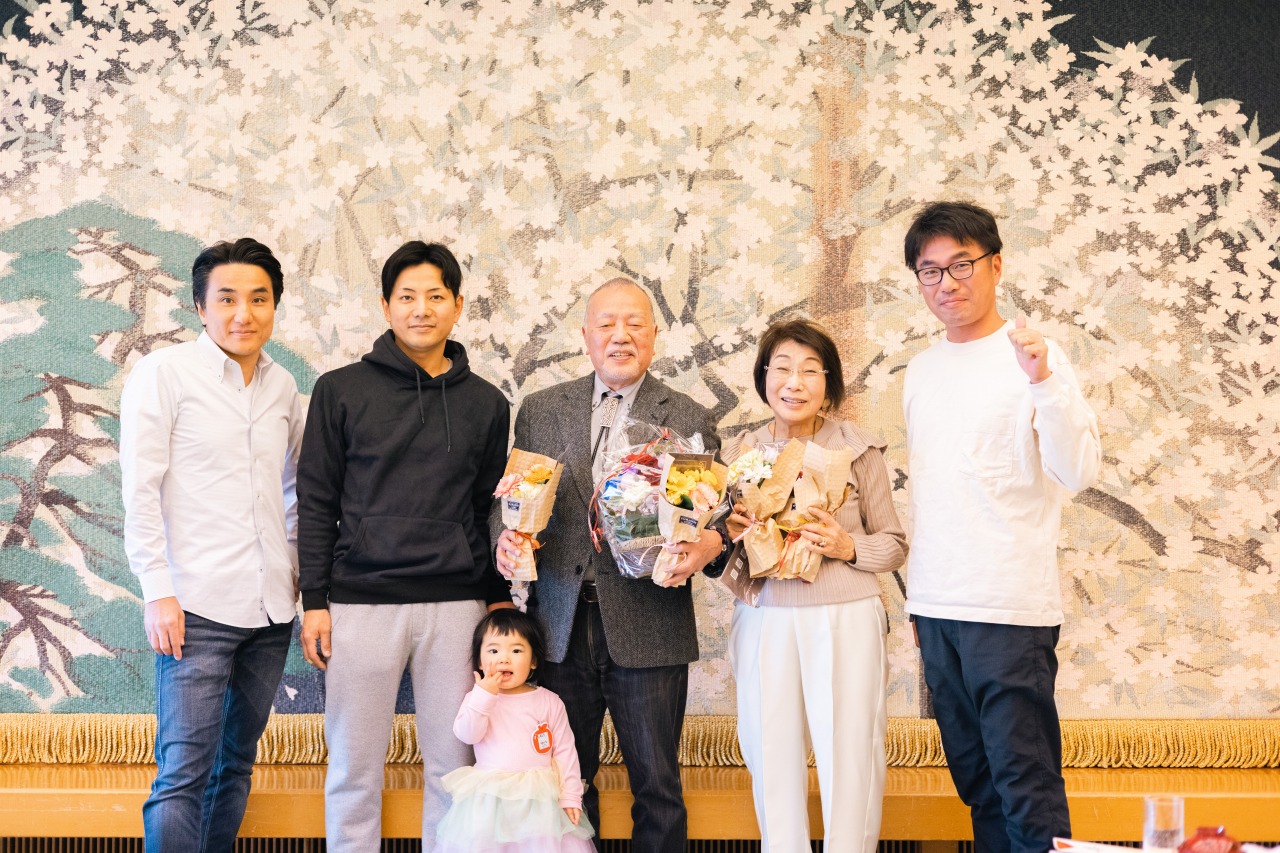 長寿祝い 家族集合写真 桜背景の会場で花束を囲む記念写真